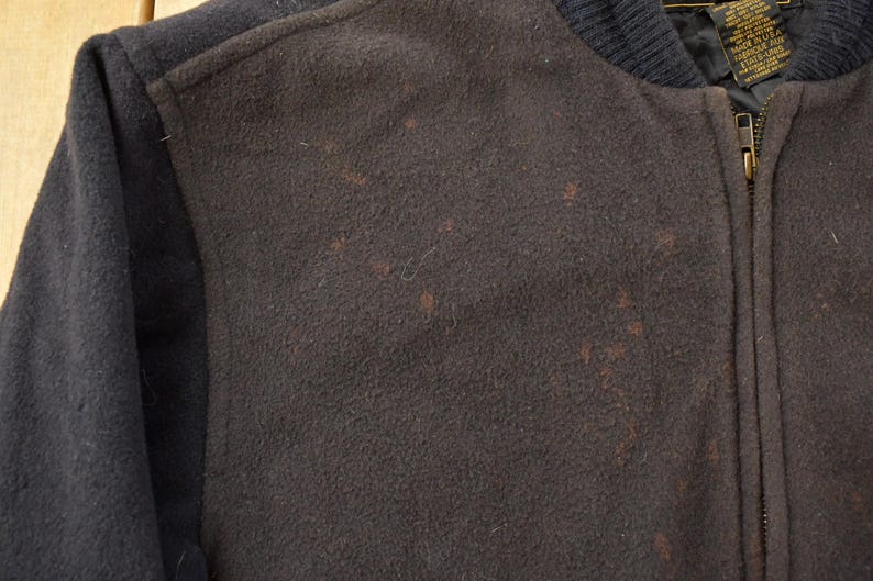 May include: A dark brown and black varsity jacket featuring a wool-like texture on the front panels and sleeves. The jacket includes a ribbed collar, a full-length front zipper, and a visible interior tag near the neckline with text in English.