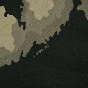 May include: A black t-shirt with a camouflage pattern in shades of brown and tan. The shirt has the text "&copy; 1995 CRI" printed on the bottom right corner.