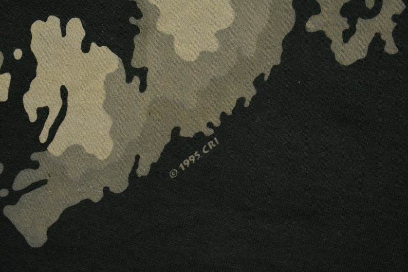 May include: A black t-shirt with a camouflage pattern in shades of brown and tan. The shirt has the text "&copy; 1995 CRI" printed on the bottom right corner.