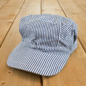 May include: Blue and white striped cap with a short brim. The cap is made of a soft fabric and has a rounded crown. The cap is sitting on a wooden surface.