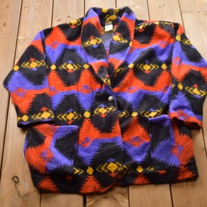 May include: A colorful fleece jacket with a geometric pattern in shades of red, blue, black, and yellow. The jacket has a shawl collar and a single button closure.