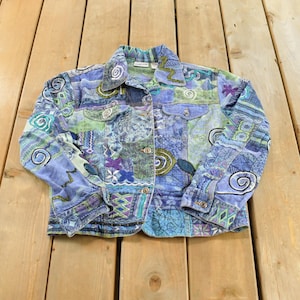 Op de afbeelding: Een spijkerjas met lange mouwen en een patchworkontwerp in verschillende tinten blauw, groen en paars. De jas is voorzien van een kraag, een knoopsluiting aan de voorzijde, twee borstzakken met klep en geborduurde spiraal- en geometrische patronen.