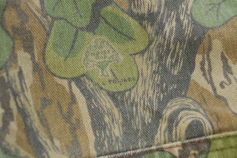 May include: Close-up of a camouflage fabric with a pattern of green, brown, and gray shapes. The word "POLAR" is visible within the design. The fabric appears to be denim or a similar sturdy material.