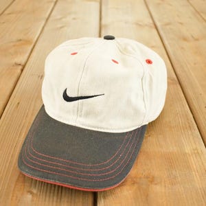 Könnte beinhalten: Eine weiße Nike Baseballkappe mit schwarzem Schirm und schwarzem Nike Swoosh-Logo. Die Kappe hat rote Nähte am Schirm und rote Akzente auf der Krone. Der Hut liegt auf einer Holzoberfläche.