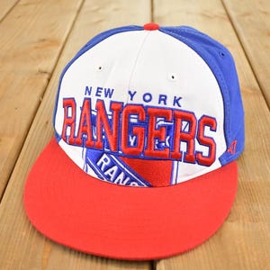 Op de afbeelding: Een New York Rangers honkbalpet met een rode klep, witte kroon en blauwe accenten. De teamnaam is rood geborduurd op de voorkant. Een klassiek sportaccessoire.