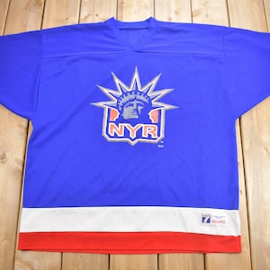 May include: A blue hockey jersey with a white and red striped collar and cuffs. The jersey features a large graphic of the Statue of Liberty with the letters "NYR" in a silver starburst. The jersey is labeled "XX-Large".