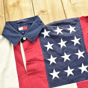 Pode incluir: Uma camisa de rugby de manga comprida vermelha, branca e azul com um logotipo Tommy Hilfiger na gola. A camisa tem um padr&atilde;o listrado e uma se&ccedil;&atilde;o azul com estrelas brancas.