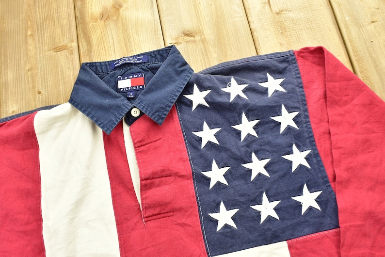 Pode incluir: Uma camisa de rugby de manga comprida vermelha, branca e azul com um logotipo Tommy Hilfiger na gola. A camisa tem um padr&atilde;o listrado e uma se&ccedil;&atilde;o azul com estrelas brancas.