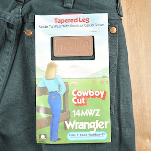 Op de afbeelding: Een groene Wrangler jeans met een label met de tekst "Tapered Leg" en "Cowboy Cut". Het label toont ook een vrouw die jeans draagt, en de tekst "14MWZ Wrangler" en "Volledige garantie van 1 jaar".