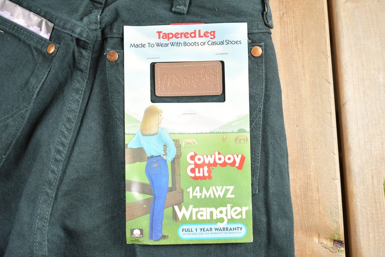 Op de afbeelding: Een groene Wrangler jeans met een label met de tekst "Tapered Leg" en "Cowboy Cut". Het label toont ook een vrouw die jeans draagt, en de tekst "14MWZ Wrangler" en "Volledige garantie van 1 jaar".