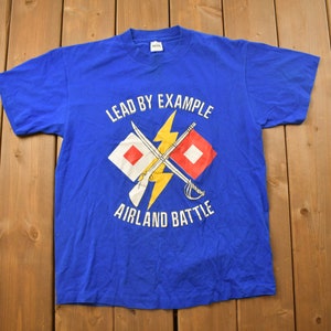 以下が含まれることがあります： 白いグラフィックデザインが施された青いTシャツ。グラフィックには、稲妻と「Lead by Example」と「Airland Battle」の文字が書かれた2つの交差した旗が描かれています。
