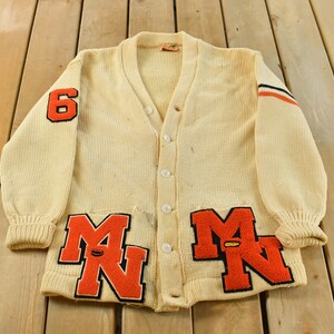 Könnte beinhalten: Cremefarbene Vintage-Strickjacke mit orangefarbenen und schwarzen Akzenten. Mit der Zahl "6" auf einem Ärmel und "MN"-Schriftzug auf der Vorderseite. Knopfverschluss und gerippte Bündchen.