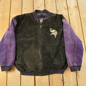 Op de afbeelding: Een vintage zwart-paarse jas met ritssluiting. De jas heeft paarse mouwen en een zwarte body met een teamlogopatch op de borst. De jas heeft een geribbelde kraag, manchetten en zoom.