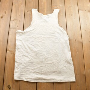May include: A white sleeveless shirt laid flat on a wooden surface.