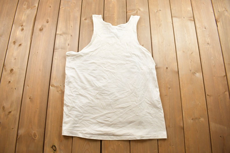 May include: A white sleeveless shirt laid flat on a wooden surface.