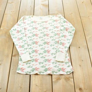 Peut inclure: Chemise thermique crème à manches longues avec un motif floral rose et vert. Le chemisier a un col rond et des poignets côtelés. Le vêtement est présenté sur une surface en bois.