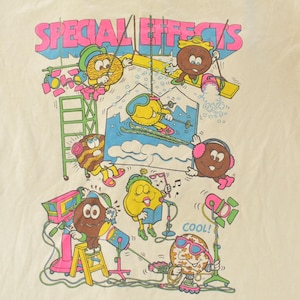 K&ouml;nnte beinhalten: Cremefarbenes T-Shirt mit einer farbenfrohen Grafik von animierten Lebensmittelcharakteren. Der Text "SPECIAL EFFECTS" steht oben in Pink und Blau. Die Charaktere sind mit verschiedenen Aktivit&auml;ten besch&auml;ftigt, darunter Skifahren und die Verwendung von B&uuml;hnenausr&uuml;stung.