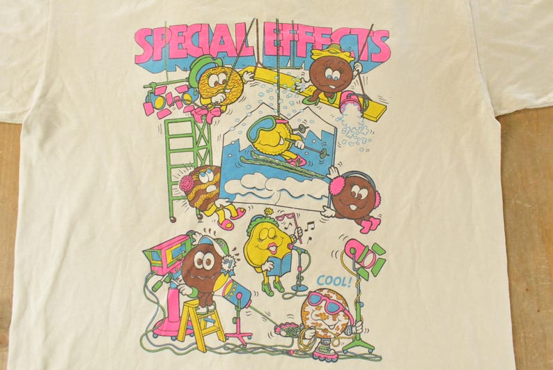 K&ouml;nnte beinhalten: Cremefarbenes T-Shirt mit einer farbenfrohen Grafik von animierten Lebensmittelcharakteren. Der Text "SPECIAL EFFECTS" steht oben in Pink und Blau. Die Charaktere sind mit verschiedenen Aktivit&auml;ten besch&auml;ftigt, darunter Skifahren und die Verwendung von B&uuml;hnenausr&uuml;stung.