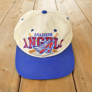 Op de afbeelding: Een witte baseballpet met een blauwe klep. De voorkant van de pet heeft een geborduurd logo met de tekst "ANAHEIM ANGELS" met een rood en wit engelenvleugelontwerp.