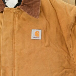 Puede incluir: Primer plano de una chaqueta de trabajo color canela con cuello de pana marr&oacute;n. La chaqueta presenta un parche cuadrado con el logotipo de Carhartt en la parte delantera. La chaqueta est&aacute; hecha de un material duradero y resistente.