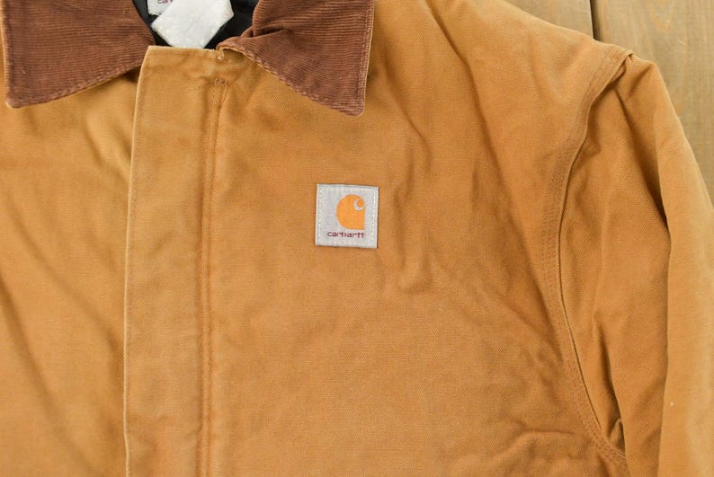 Puede incluir: Primer plano de una chaqueta de trabajo color canela con cuello de pana marr&oacute;n. La chaqueta presenta un parche cuadrado con el logotipo de Carhartt en la parte delantera. La chaqueta est&aacute; hecha de un material duradero y resistente.