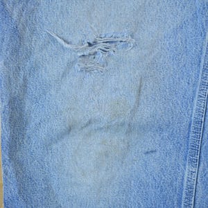 May include: Close-up of a light blue denim fabric with a small hole in the center. The fabric has a subtle texture and a faded appearance.