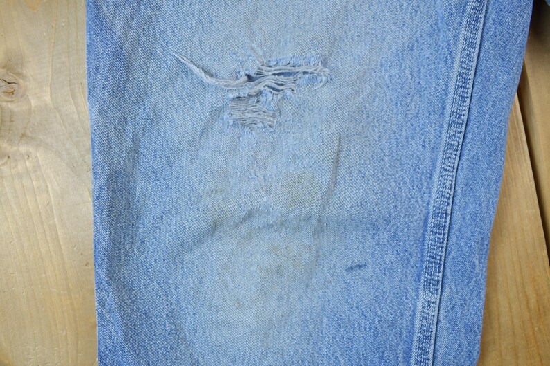 May include: Close-up of a light blue denim fabric with a small hole in the center. The fabric has a subtle texture and a faded appearance.