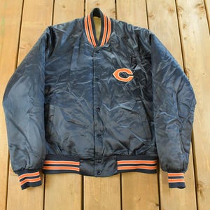 May include: Navy blue satin bomber jacket with orange and white accents. Features a button-up front, ribbed cuffs and hem, and a team logo patch on the chest. The collar is trimmed with orange and white stripes.