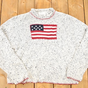 Puede incluir: Un suéter blanco moteado con un parche de la bandera estadounidense roja, blanca y azul en el pecho. El suéter tiene cuello redondo y ribete rojo en el cuello, los puños y el dobladillo. El suéter está hecho de un material de punto suave.