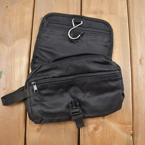 Op de afbeelding: Zwarte reistas voor toiletartikelen met een ritsvak en een haak om op te hangen. De tas is open en het interieur is zichtbaar.