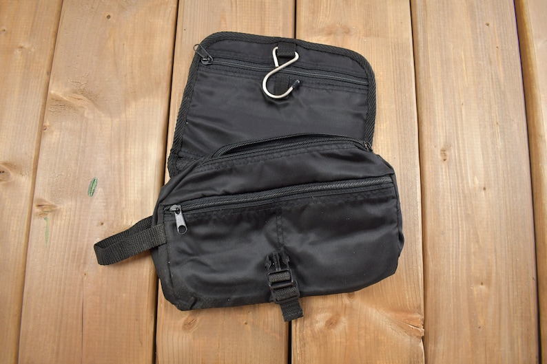 Op de afbeelding: Zwarte reistas voor toiletartikelen met een ritsvak en een haak om op te hangen. De tas is open en het interieur is zichtbaar.