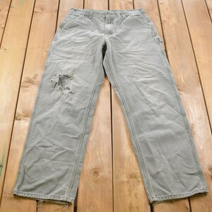 May include: A pair of grey work trousers with a distressed knee. The trousers have a straight leg and are made of a hard-wearing fabric.