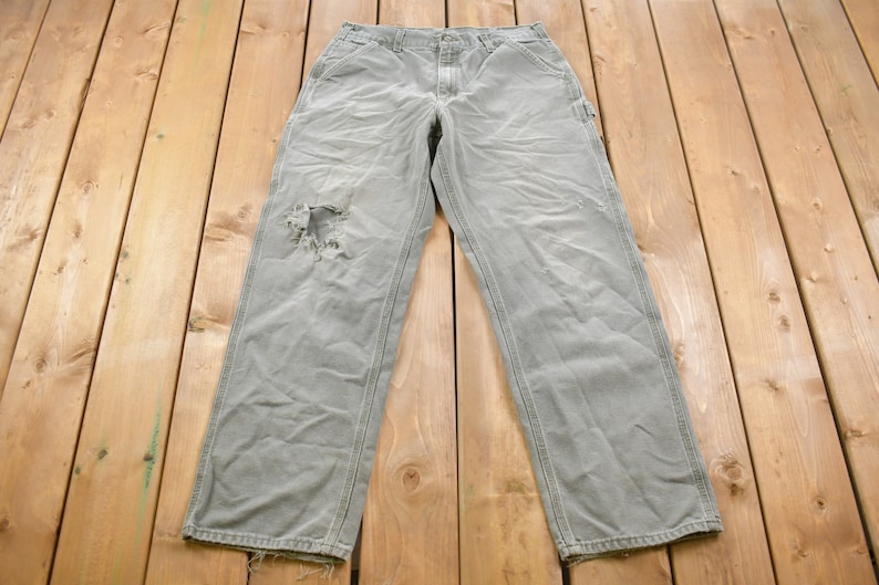 May include: A pair of grey work trousers with a distressed knee. The trousers have a straight leg and are made of a hard-wearing fabric.