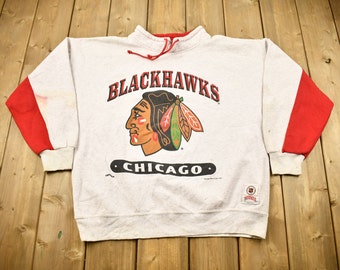 May include: A white sweatshirt with red sleeves and a black and red Chicago Blackhawks logo. The logo features a Native American head with a feather headdress and the text "Blackhawks" and "Chicago".