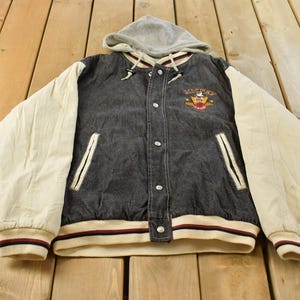 May include: A charcoal gray denim varsity jacket with cream-colored sleeves and a gray jersey hood. The jacket features a front button closure, two side pockets, and an embroidered Mickey Mouse graphic on the upper left chest with the text Mickey.