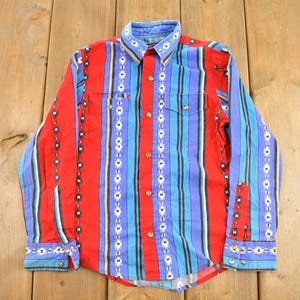 May include: A long-sleeve button-down shirt featuring a vibrant pattern of vertical red, blue, and black stripes. The shirt includes decorative white and black geometric diamond motifs along the stripes, a pointed collar, and two chest pockets.