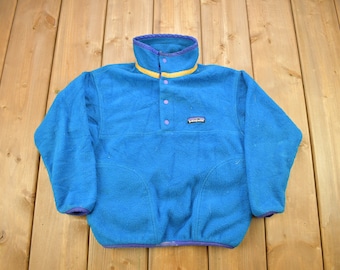 May include: A blue fleece pullover with a snap button closure, purple trim, and a yellow stripe. The Patagonia logo is visible on the left chest.