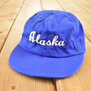 Könnte beinhalten: Blaue Baseballkappe mit dem Wort "Alaska" in weißer Schrift gestickt. Die Kappe hat einen gebogenen Schirm und besteht aus einem weichen Material. Die Kappe liegt auf einer Holzoberfläche.