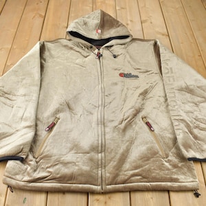 May include: A beige hooded jacket with a zipper closure and two pockets. The jacket has a quilted texture and the brand name "C.P. Company" is printed on the left chest.