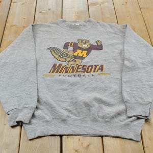 Puede incluir: Una sudadera gris jaspeado de cuello redondo con un gráfico de una ardilla de dibujos animados con una camiseta granate y dorada con la letra M. Debajo del gráfico, el texto dice MINNESOTA FOOTBALL en letras granates y doradas.