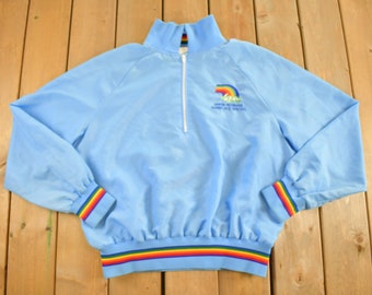 May include: A light blue quarter-zip pullover with a rainbow stripe at the bottom and cuffs. The pullover has a small embroidered logo with a rainbow and the text "SANTA MONICA RUN/WALK SPECIAL".