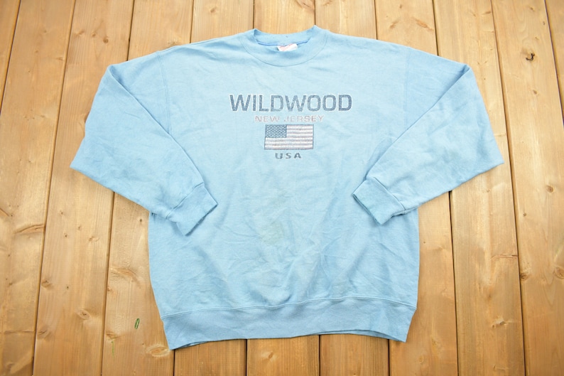 May include: A light blue sweatshirt with the text "WILDWOOD NEW JERSEY USA" and an embroidered American flag.