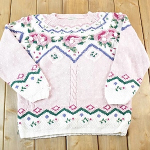 Puede incluir: Un suéter rosa claro con un diseño floral y geométrico. El suéter tiene cuello redondo y mangas largas. El diseño incluye flores rosas, hojas verdes y formas geométricas azules y verdes. El suéter está hecho de un material de punto suave.