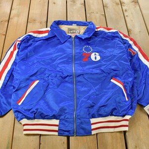 Peut inclure: Veste en satin bleu avec des rayures rouges et blanches sur les manches et les bordures. La veste a une fermeture éclair et le numéro "76" avec un cercle d'étoiles et le chiffre "6" sur la poitrine. Col et deux poches avant.