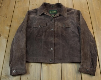 Puede incluir: Una chaqueta de ante marrón con cierre de botones y dos bolsillos de solapa. La chaqueta tiene una etiqueta que dice "DAMIER" en el cuello interior.