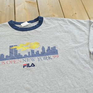 Puede incluir: Camiseta gris jaspeado con cuello y ribete de manga azul marino. Presenta un gr&aacute;fico del horizonte de Nueva York con el texto "US Open New York 99" y el logotipo de Fila. La manga tiene impreso "US Open 1999".