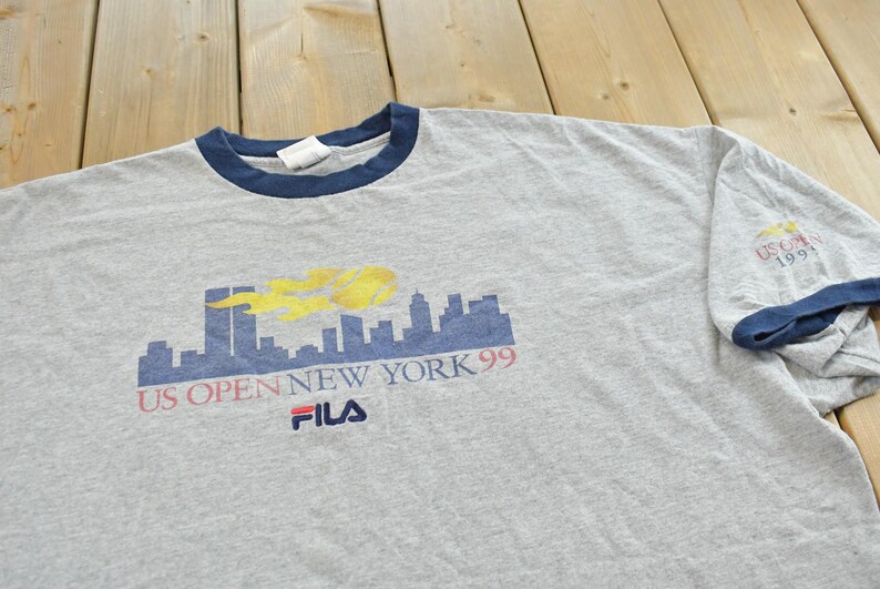 Puede incluir: Camiseta gris jaspeado con cuello y ribete de manga azul marino. Presenta un gr&aacute;fico del horizonte de Nueva York con el texto "US Open New York 99" y el logotipo de Fila. La manga tiene impreso "US Open 1999".