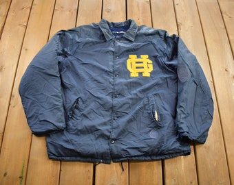 May include: Navy blue coach's jacket with a yellow "ND" logo on the chest. The jacket has a button-up front, two pockets, and a small patch on the left sleeve. The jacket is made of a lightweight material.
