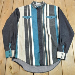 Puede incluir: Una camisa de manga larga con botones y rayas verticales en blanco, negro y azul. La camisa tiene un cuello puntiagudo y dos bolsillos en el pecho.