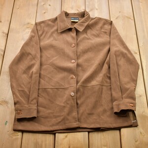 May include: A button-down, brown suede jacket with a collared neckline and long sleeves. The jacket has four buttons down the front and buttons on the cuffs. The jacket is laid flat on a light-colored wooden surface.